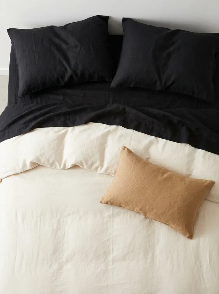 Black linen sheets layered beneath a soft cream duvet with a tan lumbar pillow